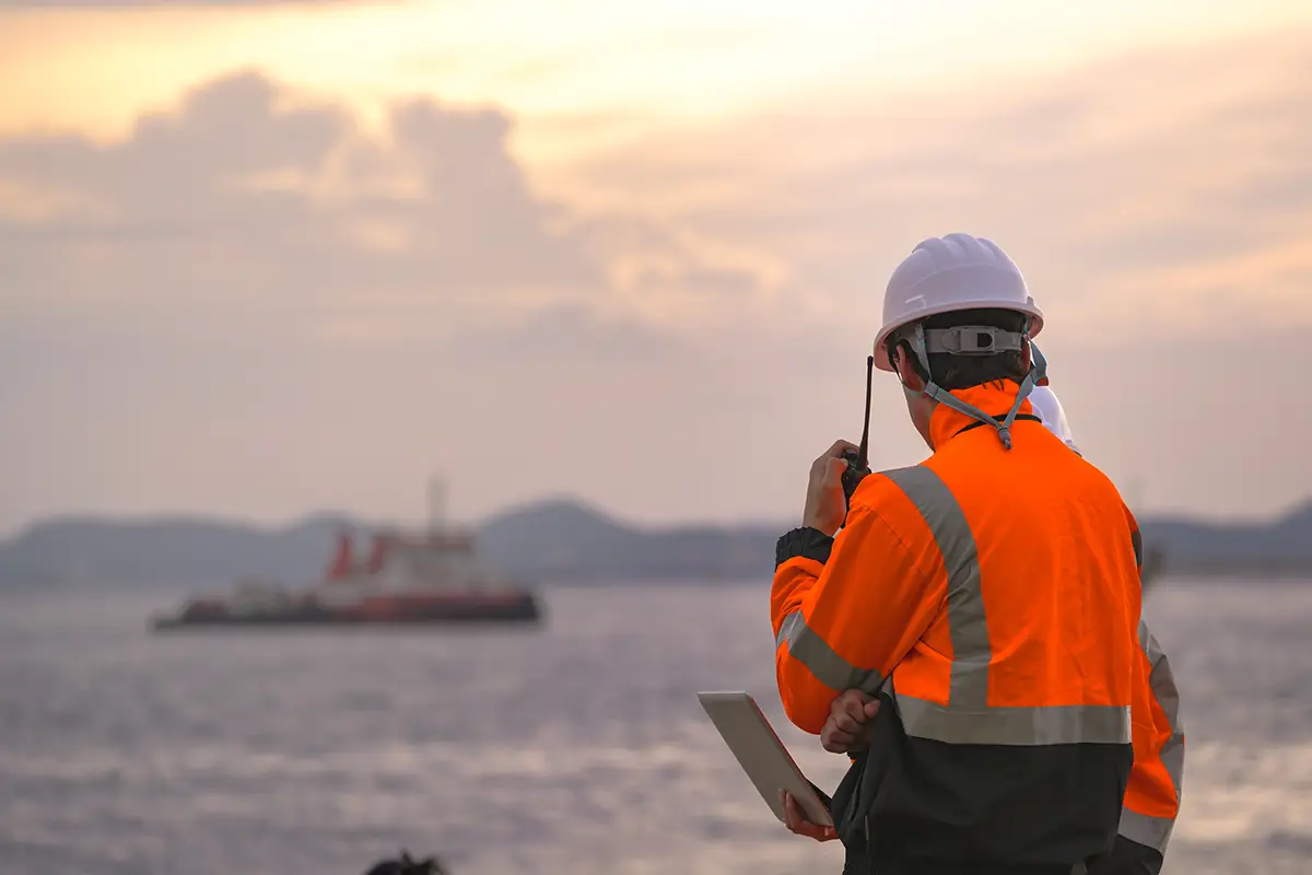 Document-Library-Img Offshore energy worker in a high-visibility orange jacket and white hard hat using a tablet or radio device, with marine vessels visible in the background at dusk